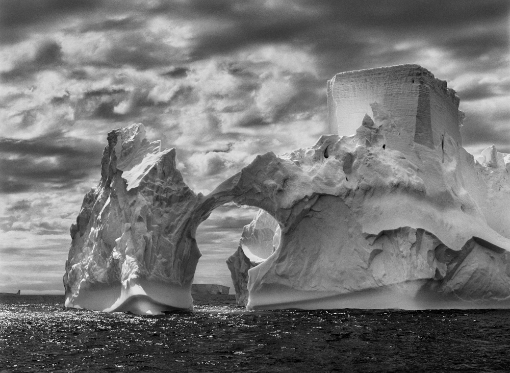 05-1-450/43.  Iceberg between Paulet Island and the South Shetland Islands on the Antarctic Channel.  At sea level, earlier flotation levels are clearly visible where the ice has been polished by the ocean’s constant movement. High above, a shape resembling a castle tower has been carved by wind erosion and detached pieces of ice.  The Antarctic Peninsula. January and February 2005.